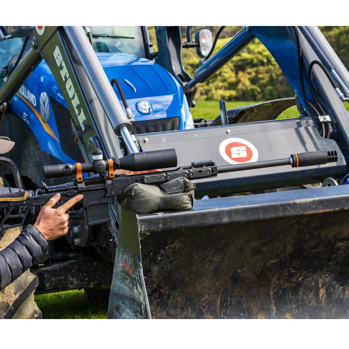 Person holding a rifle with a scope on a tractor in an outdoor setting
